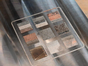 Close-up of a metal substrate with nine distinct multi-metal 3D printed alloy patches demonstrating graded compositions in metal additive manufacturing