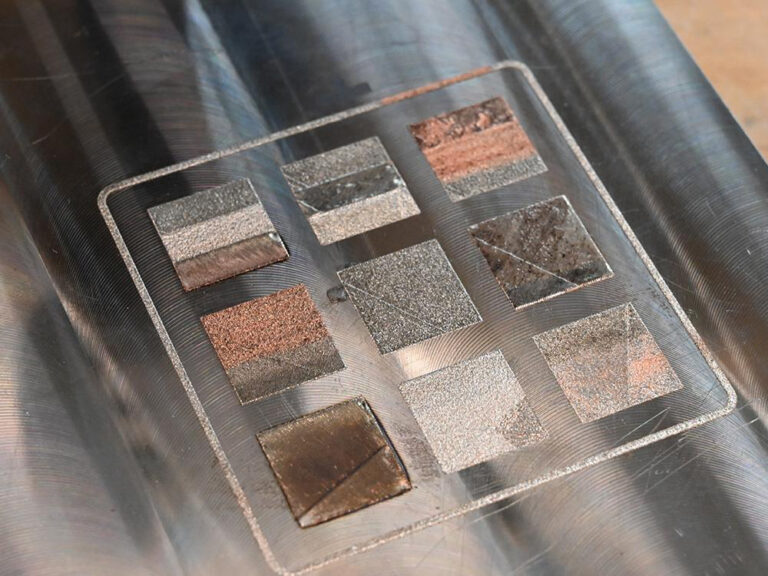 Close-up of a metal substrate with nine distinct multi-metal 3D printed alloy patches demonstrating graded compositions in metal additive manufacturing