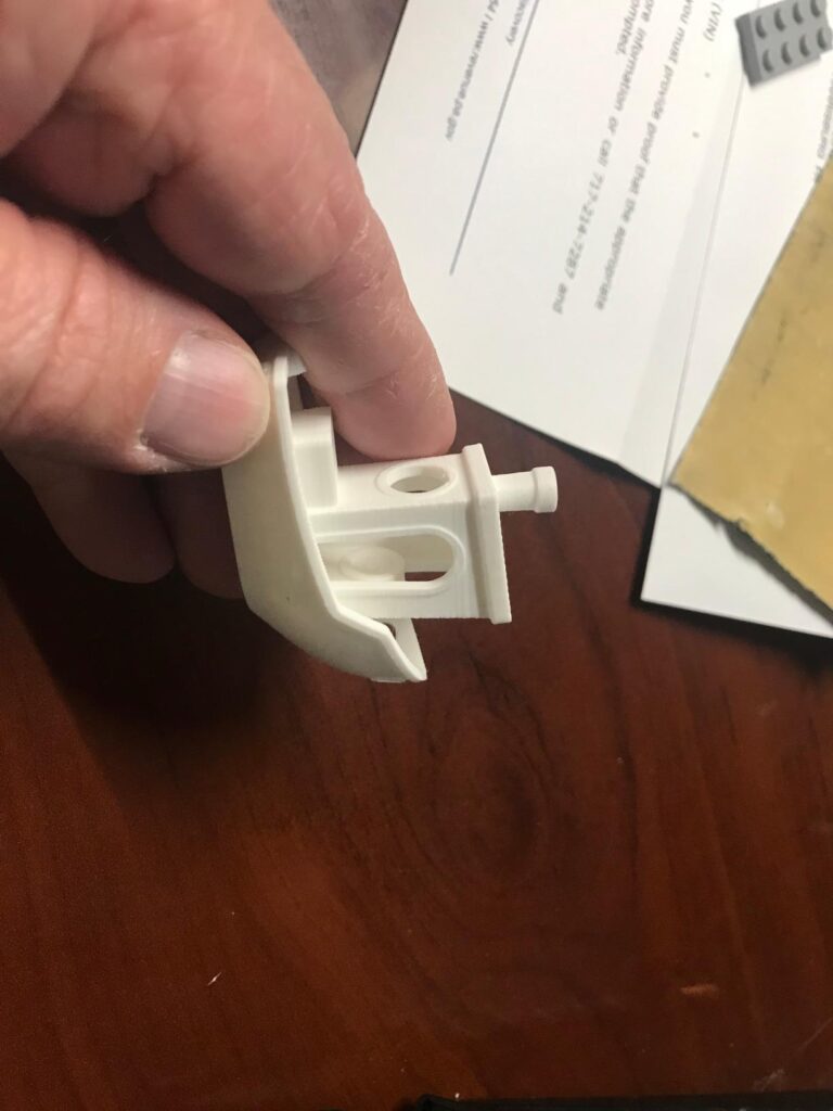 Hand holding a white bracket component 3D printed on a Creality K1C printer against a wooden desk background.