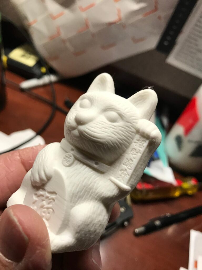 Hand holding a white Maneki-neko lucky cat figurine 3D printed with a Creality K1C, set against a cluttered desk.